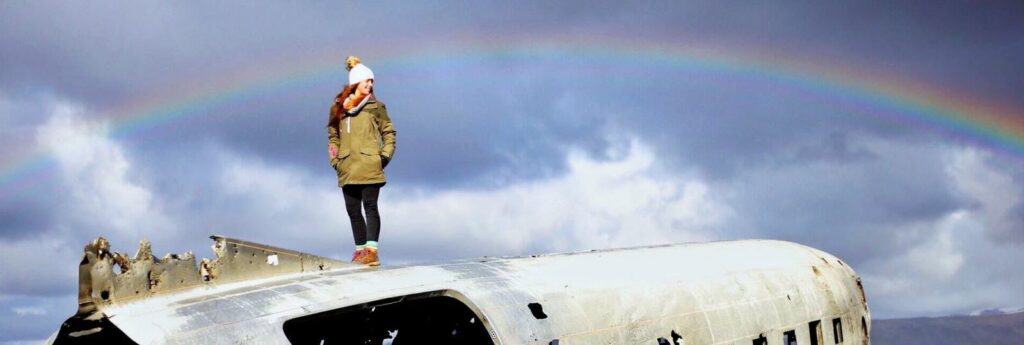 Woman standing on top of sólheimasandur plane crash with rainbow in background