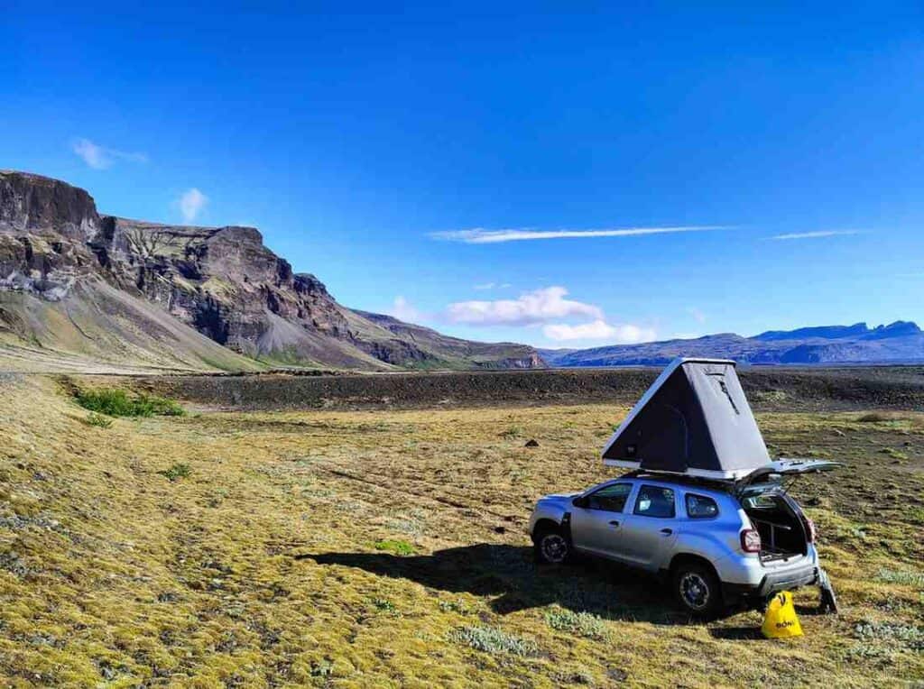 A dacia duster with an open trunk and roof tent parked by the road on grass. 
