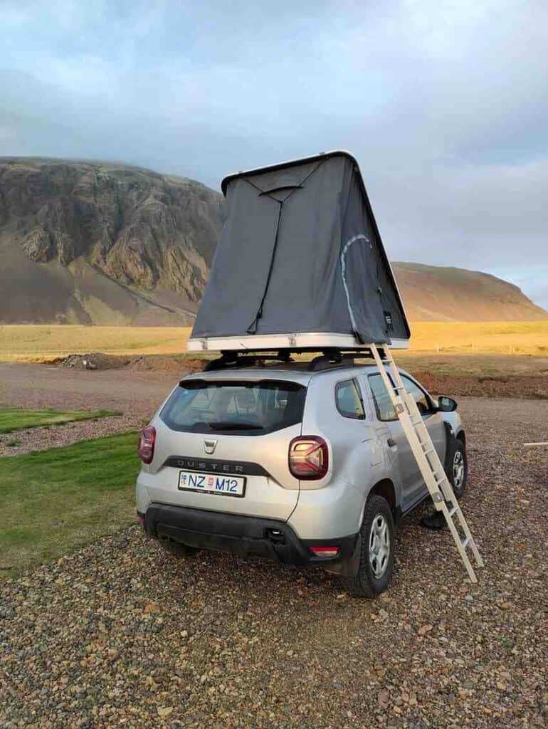 A Dacia Duster vehicle with a roof tent and a ladder placed on the side. The car is parked on a grassy field by a mountain.