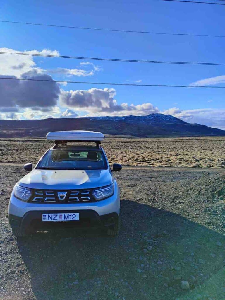 Duster parked with a closed roof tent, on a grass field, a mountain behind and blue skies.