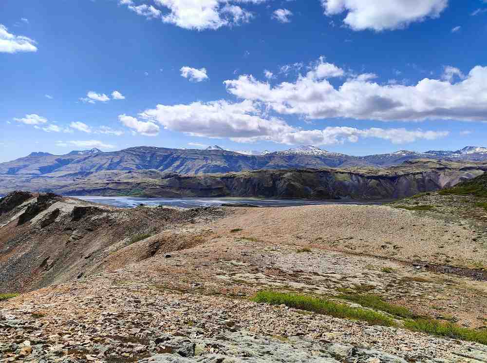 The view on top of a grassy mountain. Blue skies and a lake nearby.