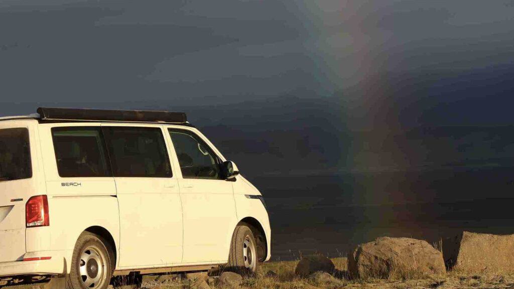 The VW California '24 parked on the edge of a cliff in the evening, a rainbow by it
