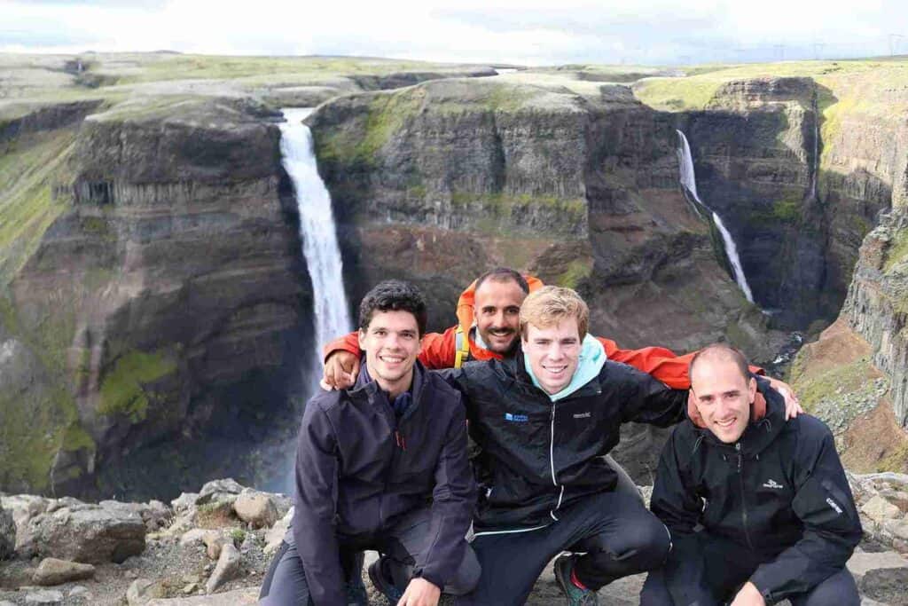 Three guys crouched by a cliffs edge, by a waterfall.