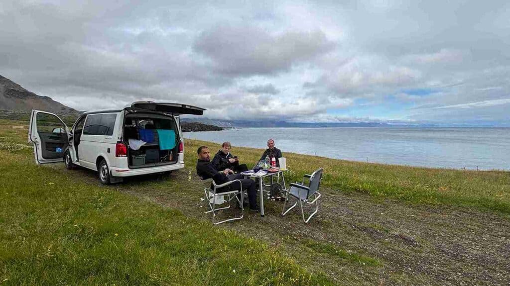 Three guys sitting on camping chairs, with a camping table, by the VW California.