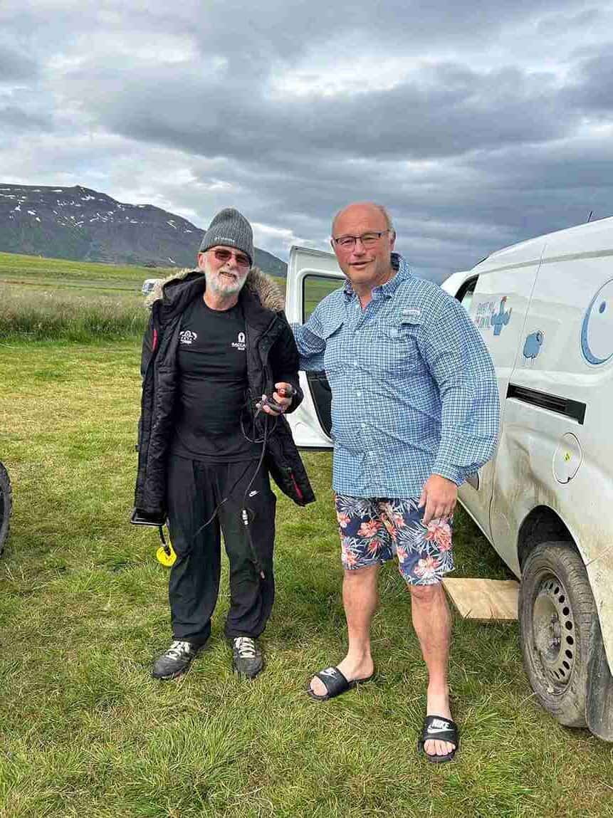 A man in shorts, button up and sandals stands with his arm around another man in joggers and a long parka jacket