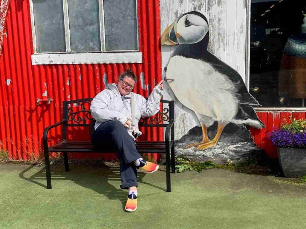 A woman sitting on a bench by a red house and a white door with a puffin painted on it.