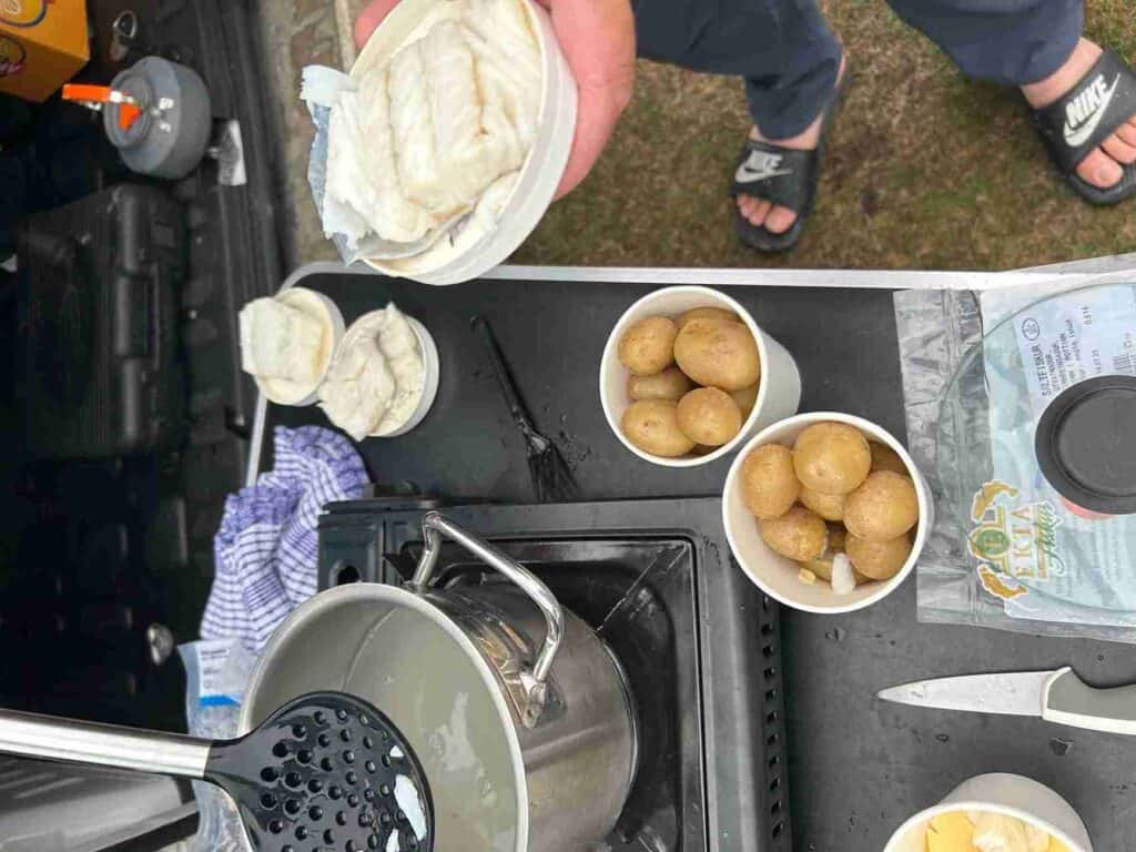 A man in sandals cooking fish and potatoes on a camping stove